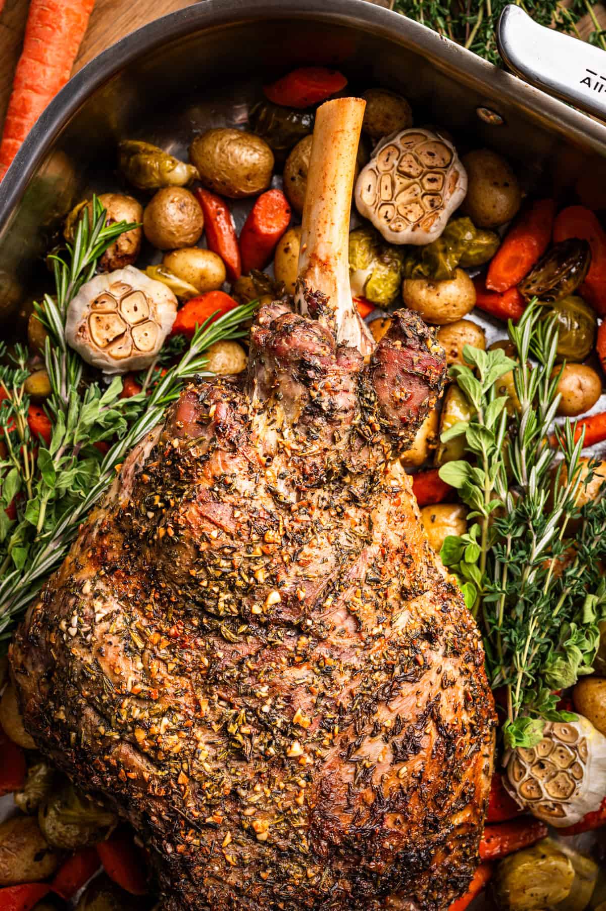 Roasted seasoned leg of lamb in a pan, surrounded by whole garlic bulbs, baby potatoes, carrots, Brussels sprouts, and fresh herbs like rosemary and oregano.