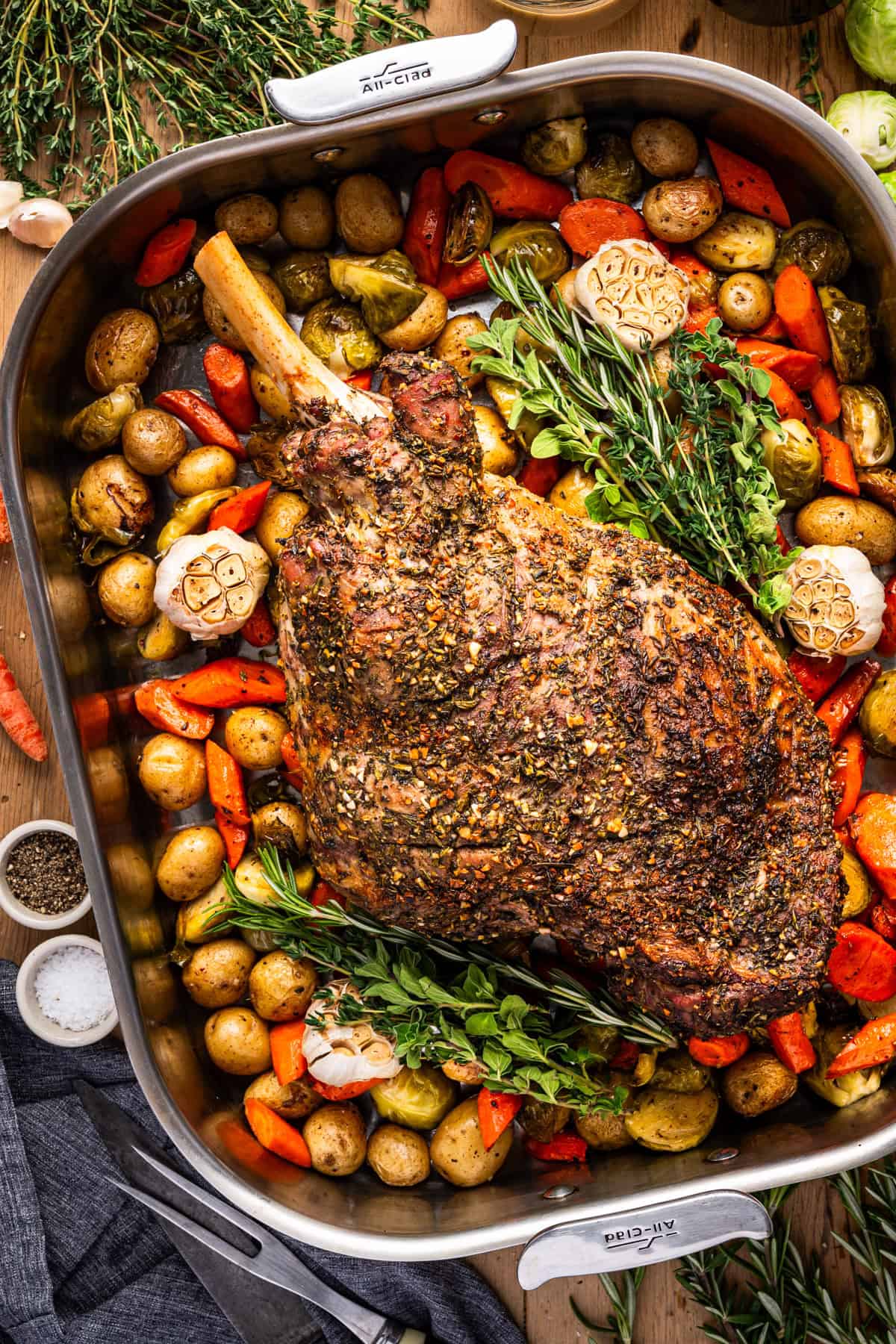 A roasted leg of lamb seasoned with herbs sits in a roasting pan surrounded by roasted baby potatoes, carrots, Brussels sprouts, whole garlic bulbs, and fresh sprigs of thyme and oregano.