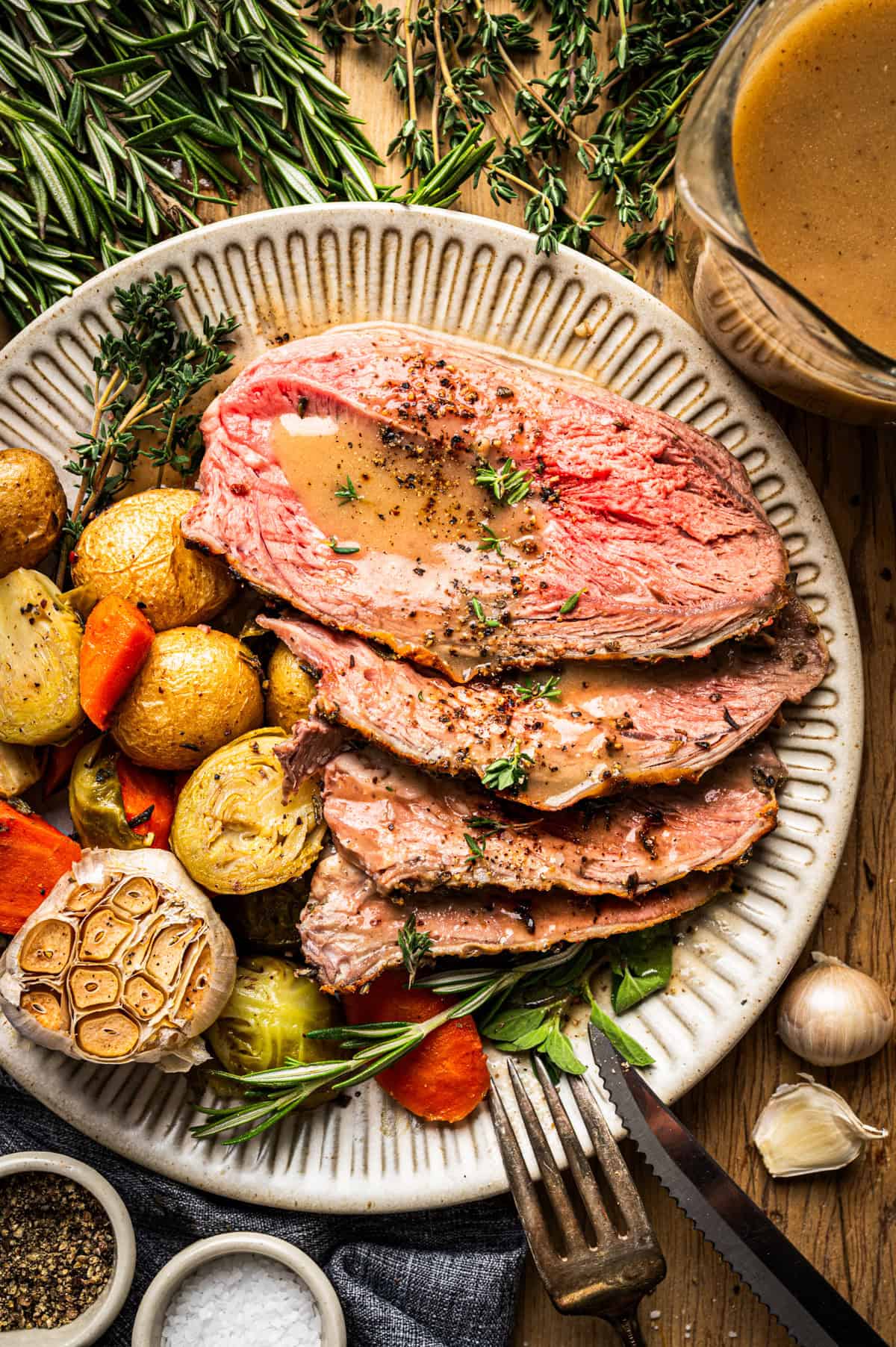 A plate with three slices of roast lamb topped with gravy, served alongside roasted baby potatoes, carrots, brussels sprouts, and a head of roasted garlic. Fresh herbs and a gravy boat are nearby.