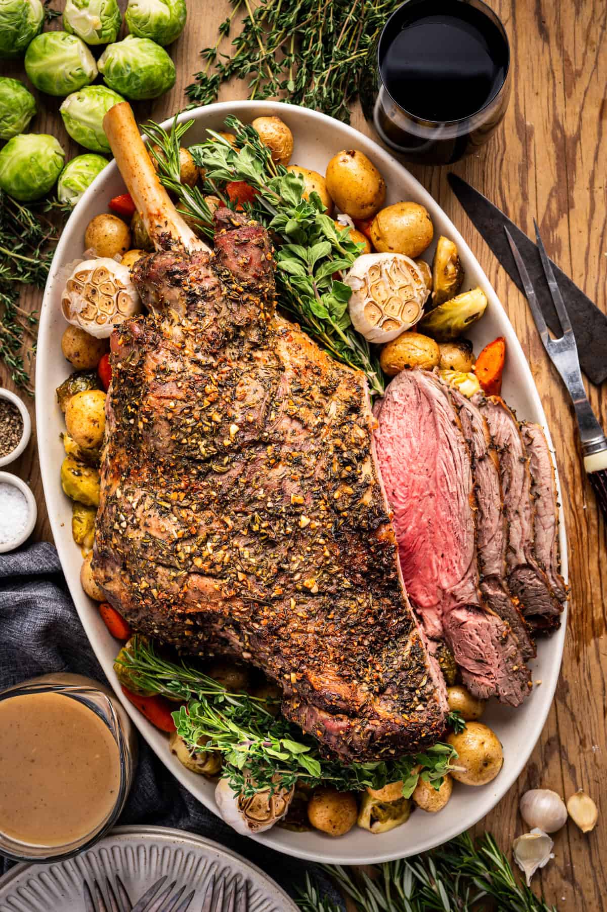 A large, seasoned bone-in roast leg of lamb is served on a platter with baby potatoes, carrots, garlic bulbs, and fresh herbs, alongside a glass of red wine and a bowl of gravy on a wooden table.