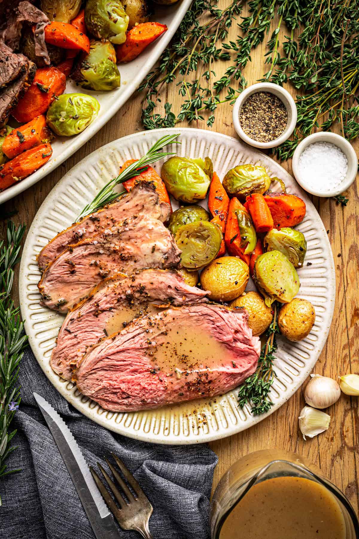 A plate with two slices of roast lamb topped with gravy, served with roasted potatoes, carrots, Brussels sprouts, and fresh herbs. Salt, pepper, and a platter of more vegetables are nearby on a wooden table.