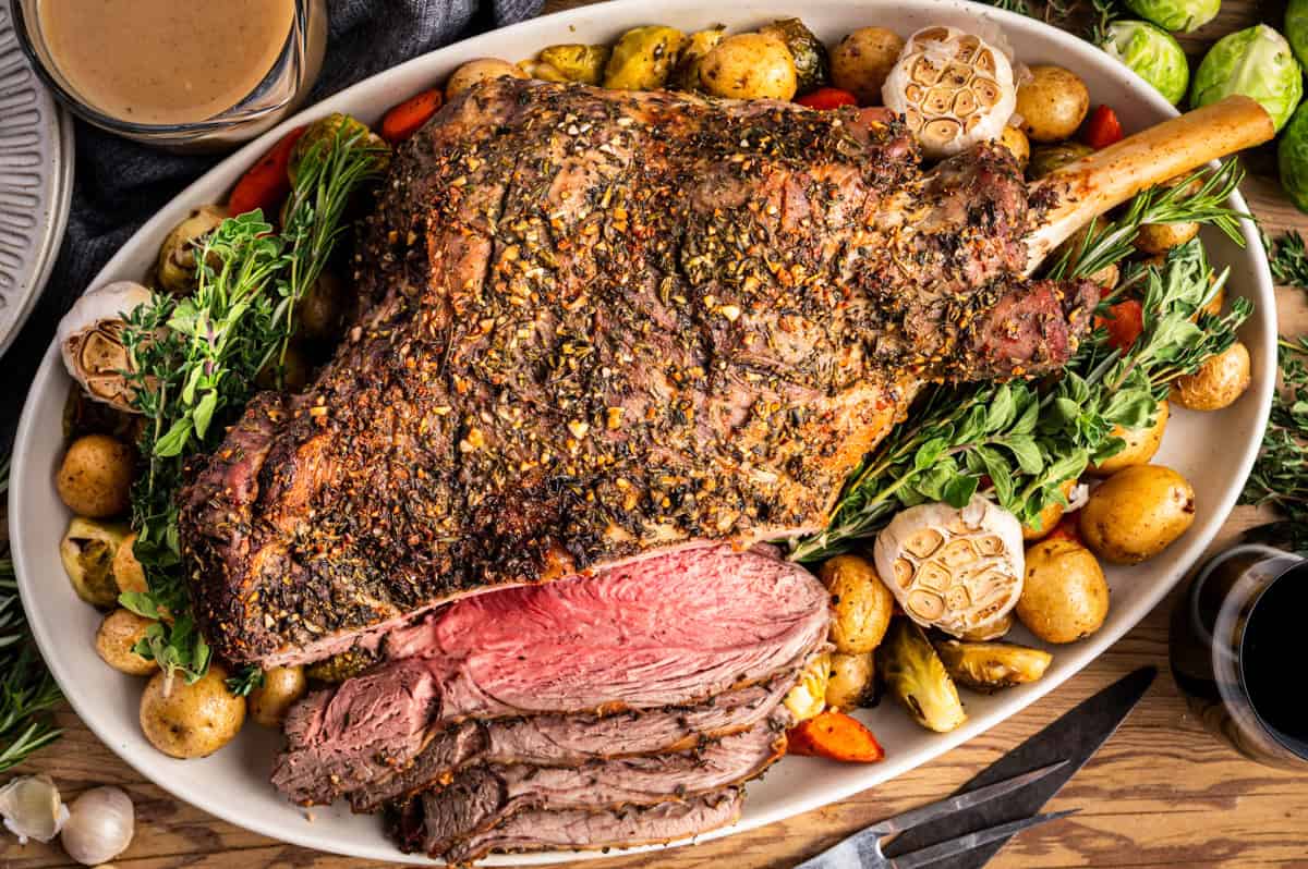A roasted leg of lamb garnished with herbs, surrounded by roasted potatoes, carrots, and whole garlic bulbs on a platter. Several slices of the lamb are cut to reveal the juicy pink interior.