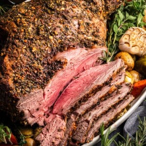 A close-up of a herb-crusted roasted leg of lamb, sliced to reveal juicy pink meat inside, surrounded by roasted potatoes, carrots, fresh herbs, and a halved head of garlic.