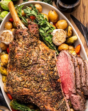 A large roasted leg of lamb, seasoned with herbs, is served on a platter with baby potatoes, carrots, Brussels sprouts, whole garlic bulbs, and fresh herbs, with several slices carved and a glass of red wine nearby.