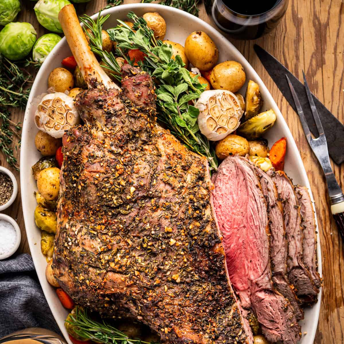 A large roasted leg of lamb, seasoned with herbs, is served on a platter with baby potatoes, carrots, Brussels sprouts, whole garlic bulbs, and fresh herbs, with several slices carved and a glass of red wine nearby.