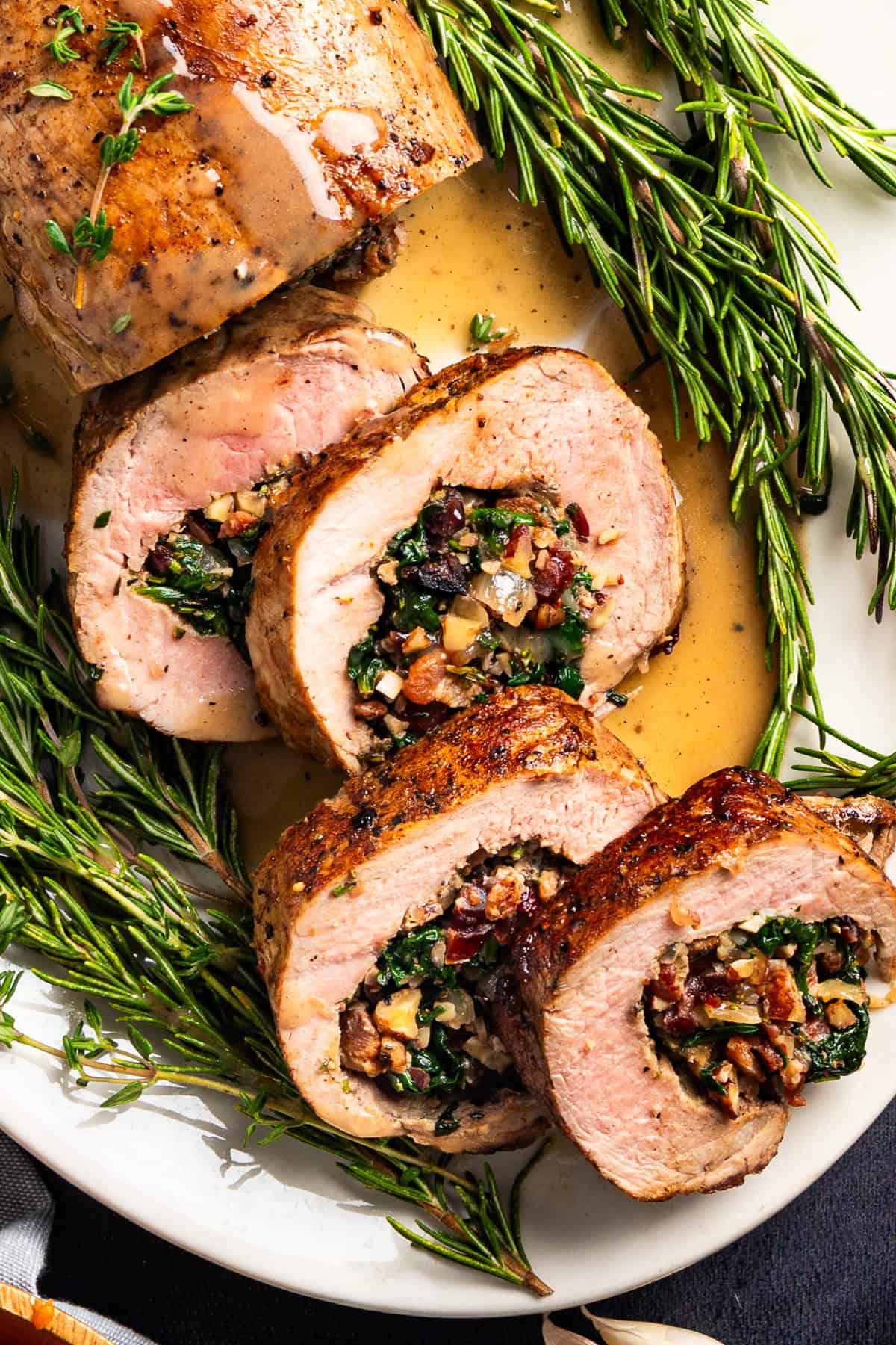 Pork Tenderloin Stuffed with Spinach, Bacon, and Dried Cranberries Sliced stuffed pork tenderloin filled with spinach, pecans, bacon, dried cranberries, and herbs, arranged on a white plate with sprigs of fresh rosemary and a light sauce.