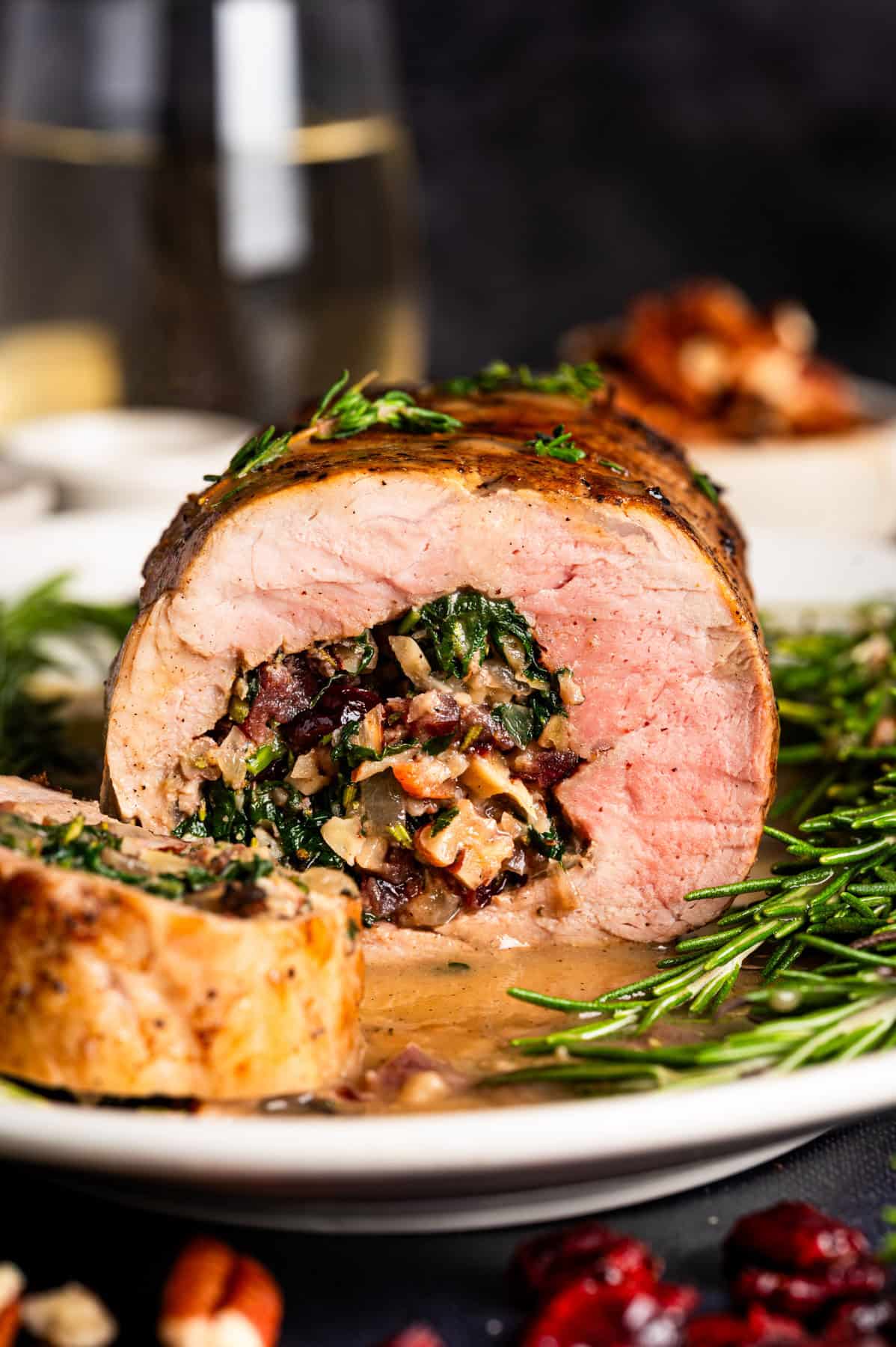 Pork Tenderloin Stuffed with Spinach, Bacon, and Dried Cranberries A sliced pork tenderloin stuffed with spinach, bacon, pecans, dried cranberries, and herbs, served with creamy sauce and garnished with fresh rosemary on a white plate.