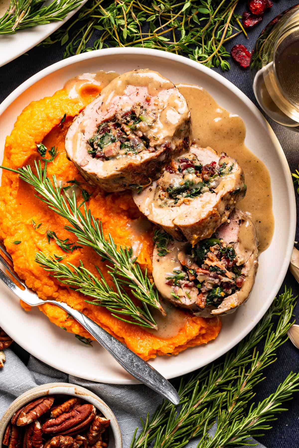Pork Tenderloin Stuffed with Spinach, Bacon, and Dried Cranberries A plate with three slices of stuffed pork roulade topped with gravy, served alongside a generous portion of mashed sweet potatoes and garnished with fresh rosemary sprigs.