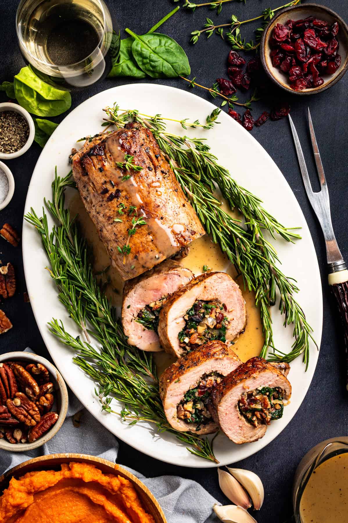 Pork Tenderloin Stuffed with Spinach, Bacon, and Dried Cranberries A white platter with herb-crusted pork tenderloin, partially sliced to reveal a stuffing of greens, bacon, pecans, and dried fruit, garnished with rosemary. Surrounding sides include mashed sweet potatoes, nuts, cranberries, and a glass of white wine.
