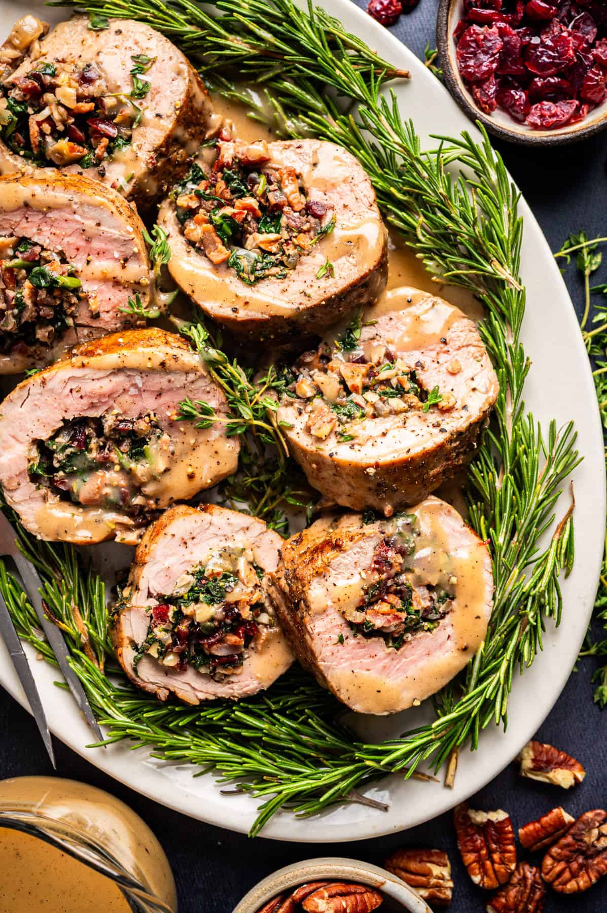 Pork Tenderloin Stuffed with Spinach, Bacon, and Dried Cranberries Sliced stuffed pork tenderloin with herbs, spinach, nuts, and cranberries, arranged on a white platter with fresh rosemary sprigs, served with gravy. Small bowls of cranberries and pecans are nearby.