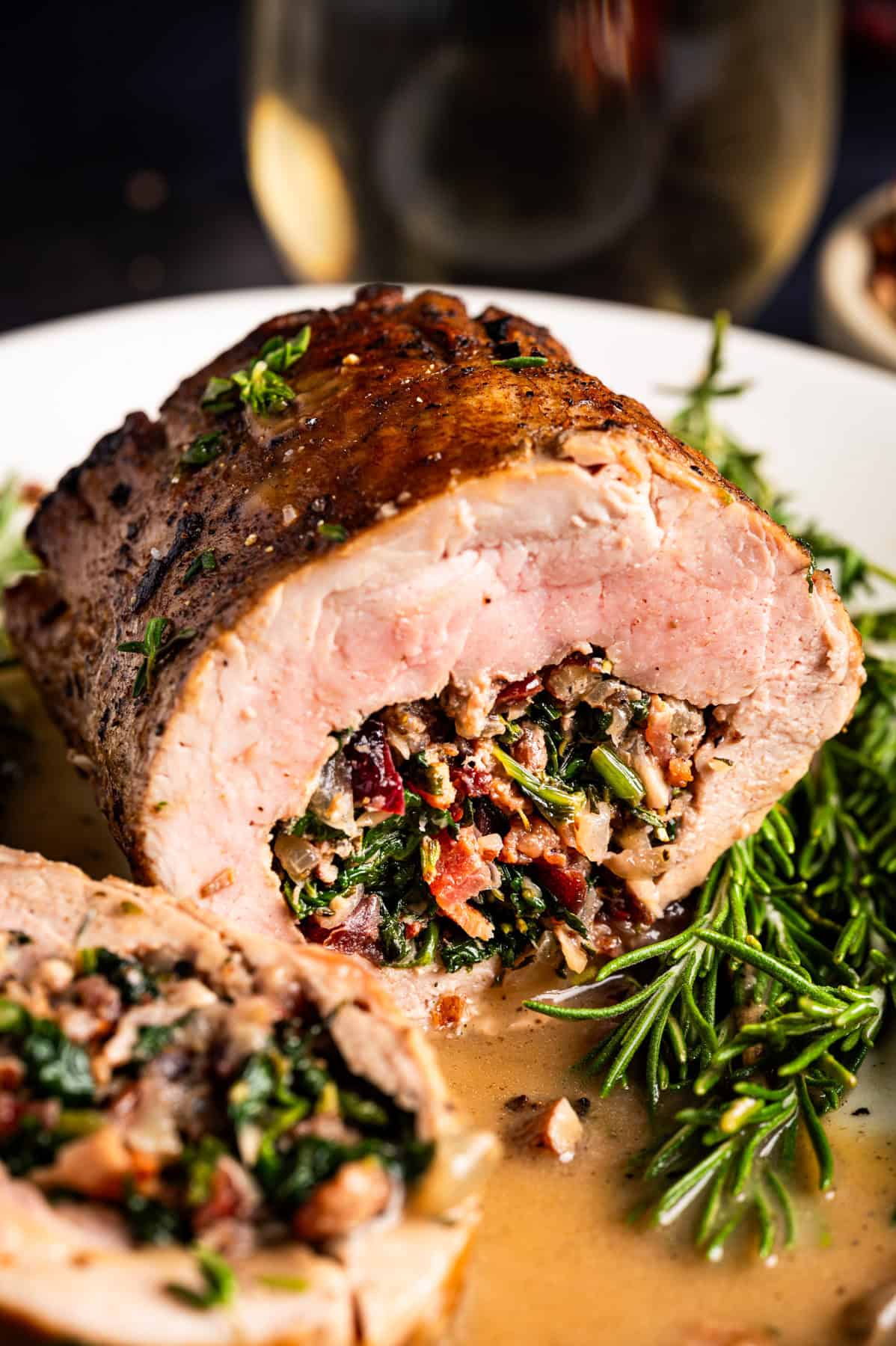 Pork Tenderloin Stuffed with Spinach, Bacon, and Dried Cranberries A close-up of a sliced stuffed pork tenderloin, filled with spinach, bacon, dried cranberries, pecans, and herbs, served with sauce and garnished with fresh rosemary on a white plate.