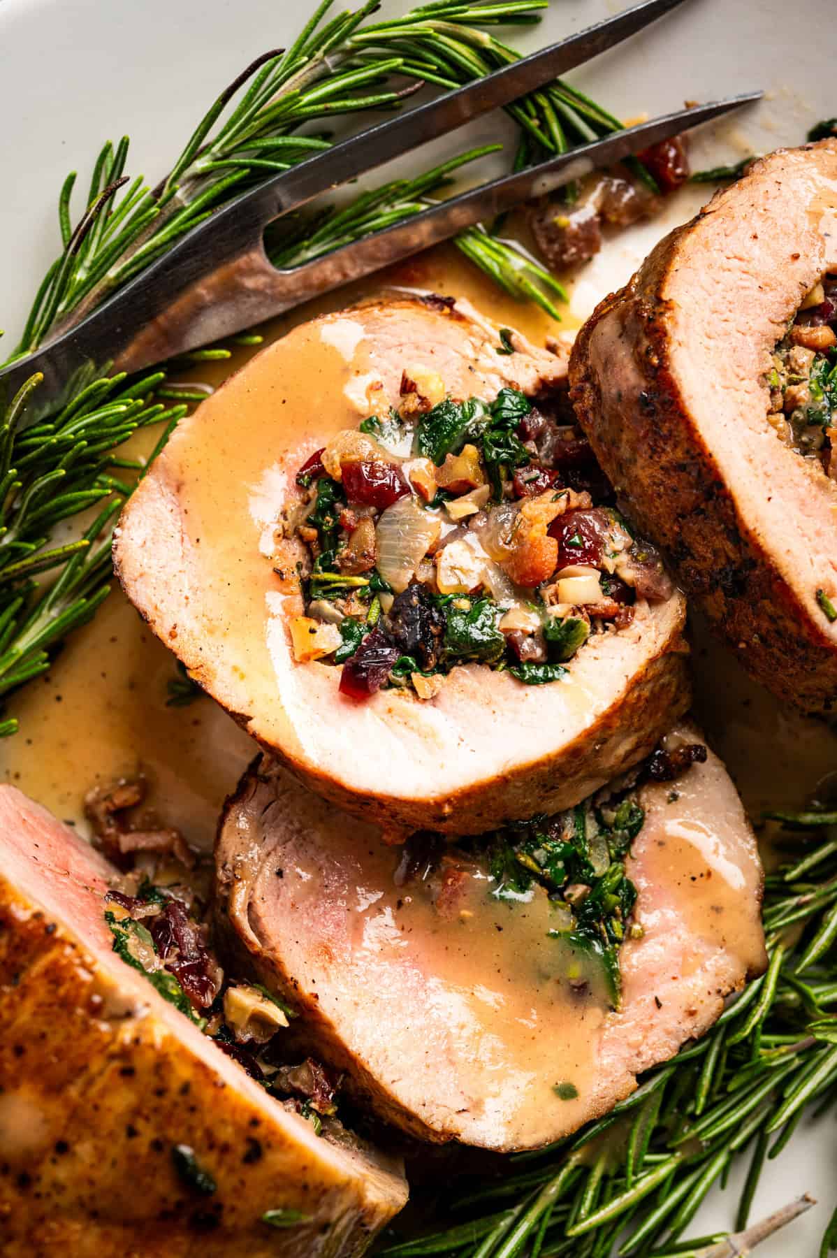 Pork Tenderloin Stuffed with Spinach, Bacon, and Dried Cranberries Close-up of sliced stuffed pork tenderloin with a savory filling of spinach, herbs, dried cranberries, and nuts, served with gravy and garnished with fresh rosemary sprigs and a metal serving fork.