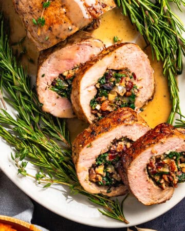 Sliced stuffed pork tenderloin with a filling of spinach, pecans, dried cranberries, bacon, and herbs, served on a white platter garnished with fresh rosemary and drizzled with sauce.