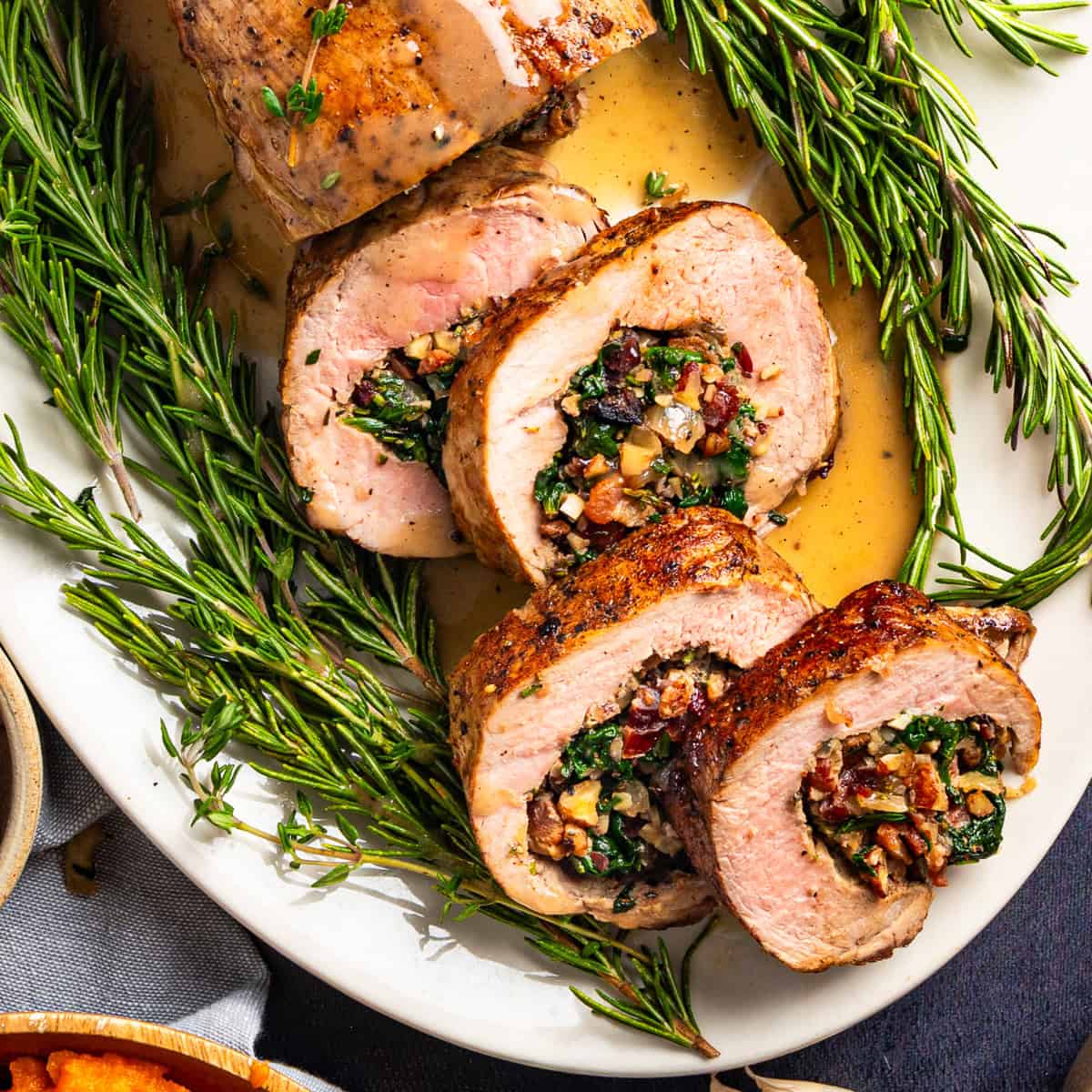 Sliced stuffed pork tenderloin with a filling of spinach, pecans, dried cranberries, bacon, and herbs, served on a white platter garnished with fresh rosemary and drizzled with sauce.