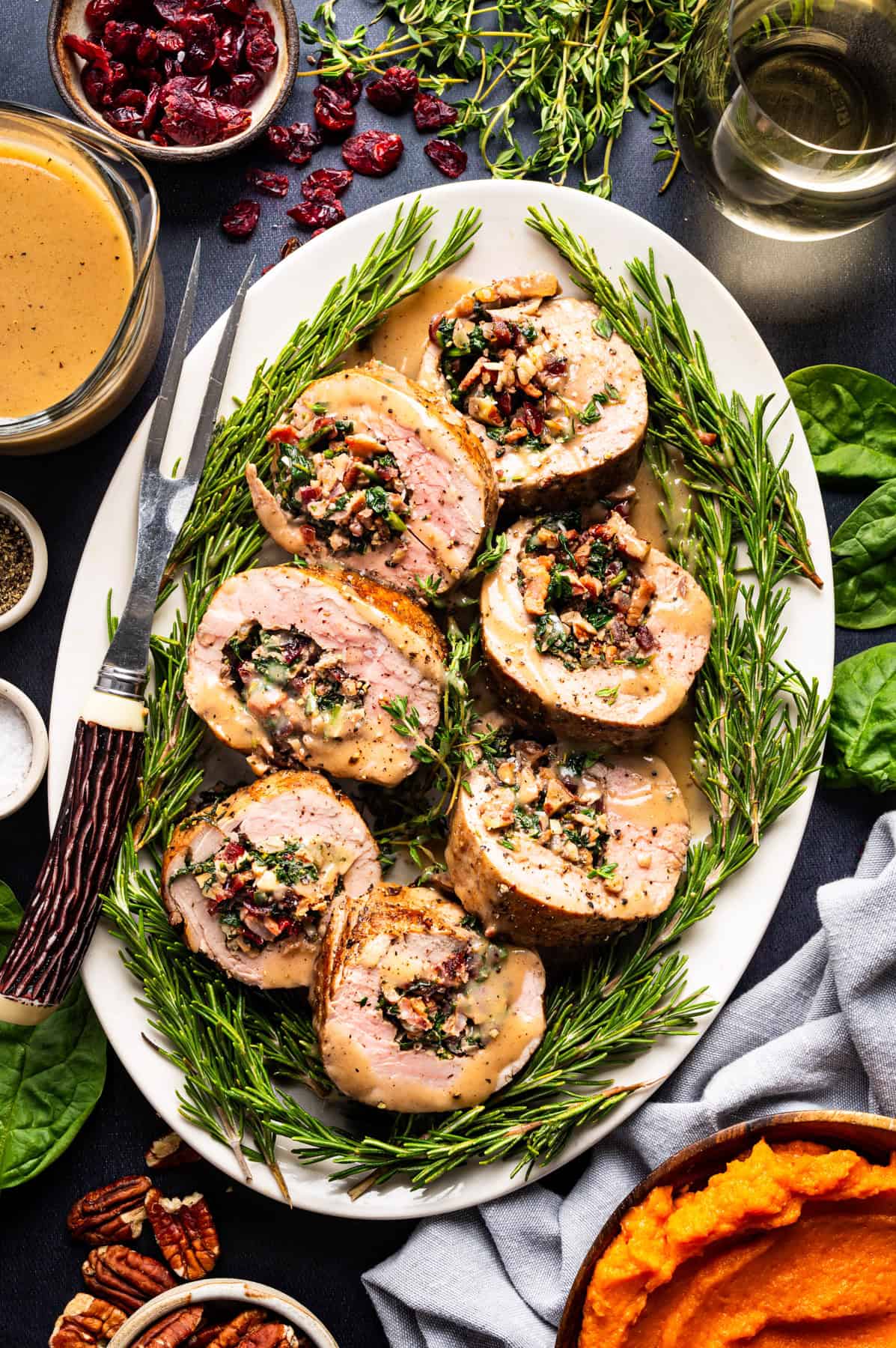 Pork Tenderloin Stuffed with Spinach, Bacon, and Dried Cranberries Sliced stuffed pork tenderloin with creamy sauce, garnished with fresh rosemary on a white platter, surrounded by pecans, cranberries, greens, and a bowl of mashed sweet potatoes. A carving fork and sauce are beside the platter.