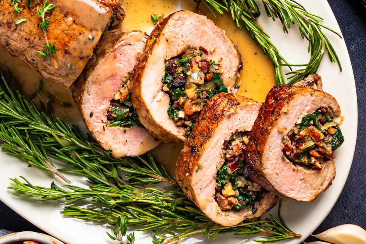 This Stuffed Pork Tenderloin is a savory-sweet main dish made by butterflying and filling pork tenderloin with crispy bacon, sautéed spinach, fresh herbs, dried cranberries, and toasted pecans, then roasting it until juicy and perfectly cooked (perfect for a weeknight dinner or holiday main course). Sliced stuffed pork tenderloin on a white plate, filled with spinach, pecans, bacon, and dried cranberries, surrounded by sprigs of fresh rosemary and drizzled with a light sauce.
