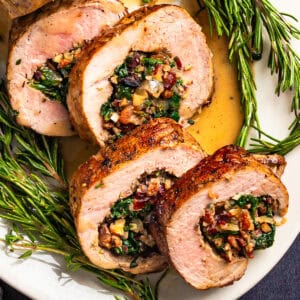 Sliced stuffed pork tenderloin with a savory filling of spinach, bacon, dried cranberries, pecans, garnished with fresh rosemary sprigs and served with a light sauce on a white plate.