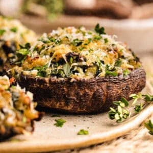 A close-up of a stuffed portobello mushroom topped with melted cheese, spinach, herbs, and breadcrumbs, served on a rustic ceramic plate with fresh chopped parsley sprinkled around.