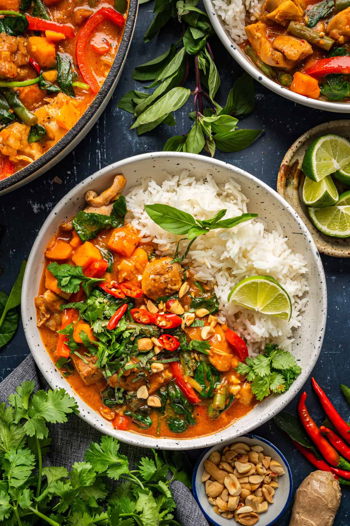 Thai Chicken Curry A bowl of Thai red curry with chicken, bell peppers, and greens, served with steamed white rice, lime wedge, fresh herbs, chopped peanuts, and sliced red chilies. Additional herbs and sides are nearby on a dark surface.