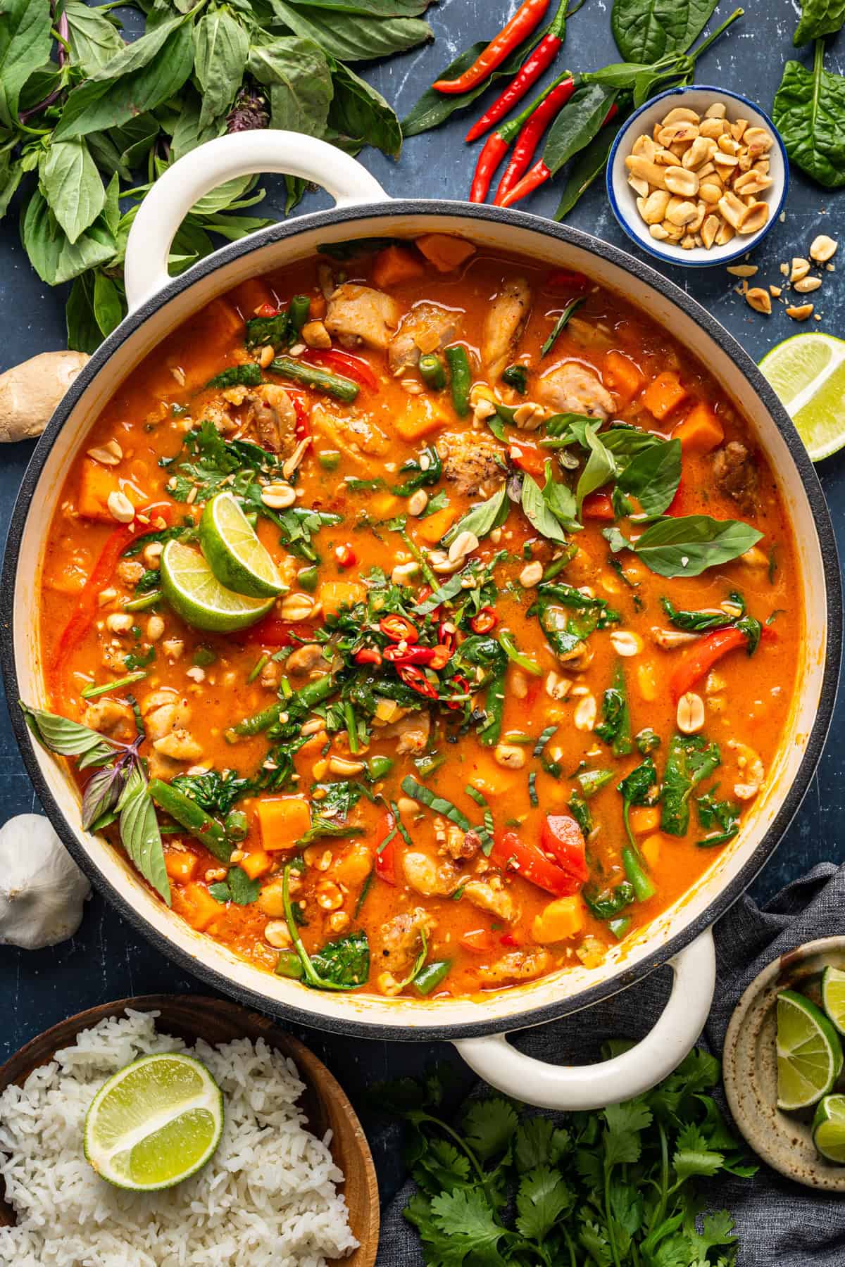 Thai Chicken Curry A pot of vibrant Thai chicken red curry with vegetables, topped with fresh basil, lime wedges, chopped peanuts, and chilies. Surrounded by rice, herbs, lime, chili peppers, peanuts, and garlic on a dark surface.