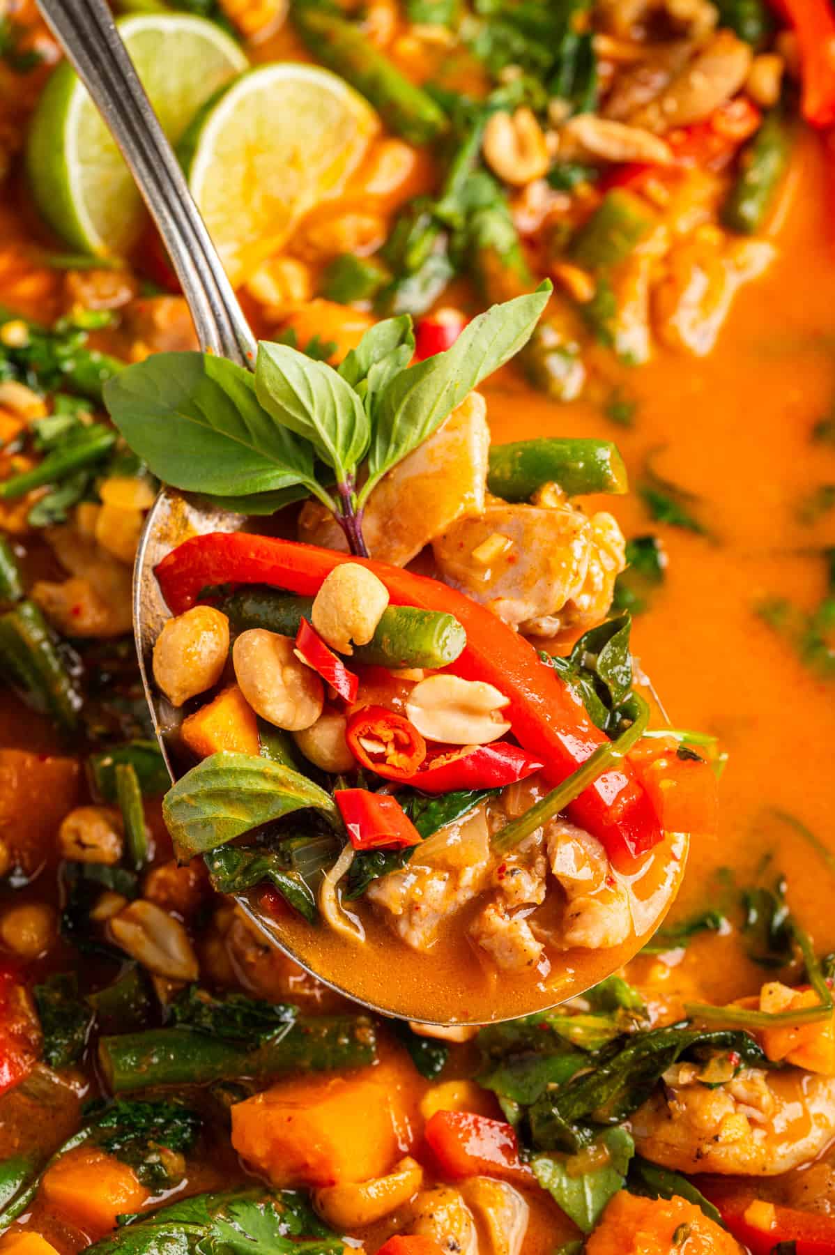 Thai Chicken Curry A close-up of a spoonful of Thai red curry with chicken, red bell peppers, green beans, peanuts, fresh basil, and red chili slices in a creamy orange sauce, with lime wedges visible in the background.