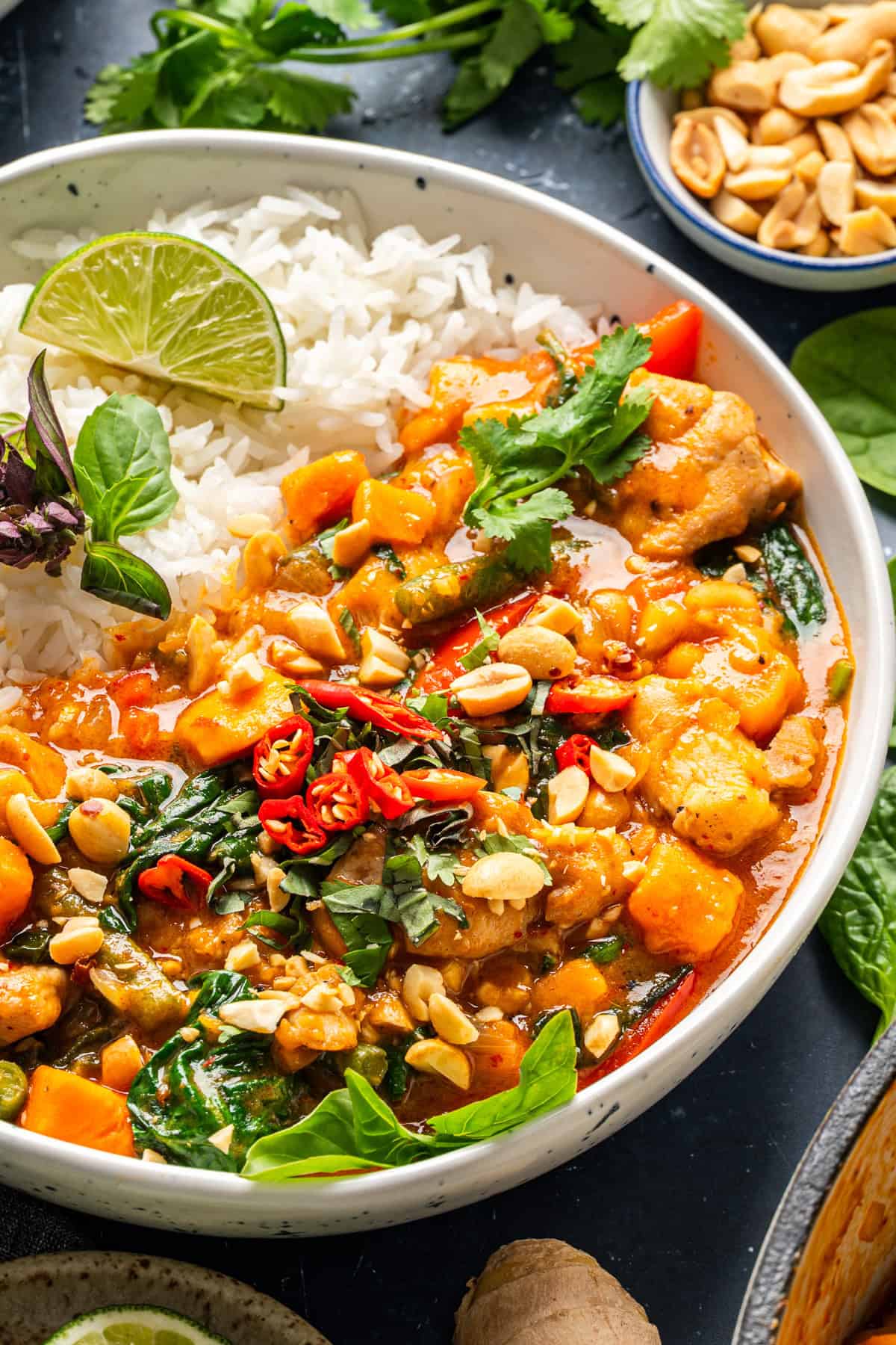 Thai Chicken Curry A bowl of white rice served with a colorful Thai curry containing chicken, vegetables, spinach, chopped red chilies, fresh herbs, lime, and sprinkled with peanuts. A small bowl of peanuts is visible in the background.