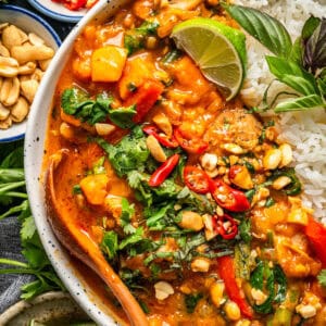 A bowl of vibrant Thai chicken curry with vegetables, topped with fresh cilantro, sliced red chili, peanuts, and a lime wedge, served with white rice and a wooden spoon.