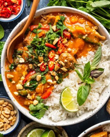 A bowl of Thai red curry with chicken, vegetables and peanuts, served alongside white rice, fresh herbs, sliced red chili, and a lime wedge, surrounded by bowls of peanuts, greens, and chili on a dark surface.