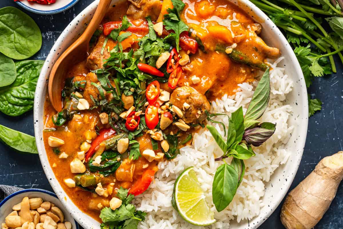 Thai Chicken Curry A bowl of white rice served with Thai red curry with chicken, topped with fresh herbs, chopped peanuts, sliced red chili, and a lime wedge, garnished with basil and surrounded by fresh spinach and cilantro.