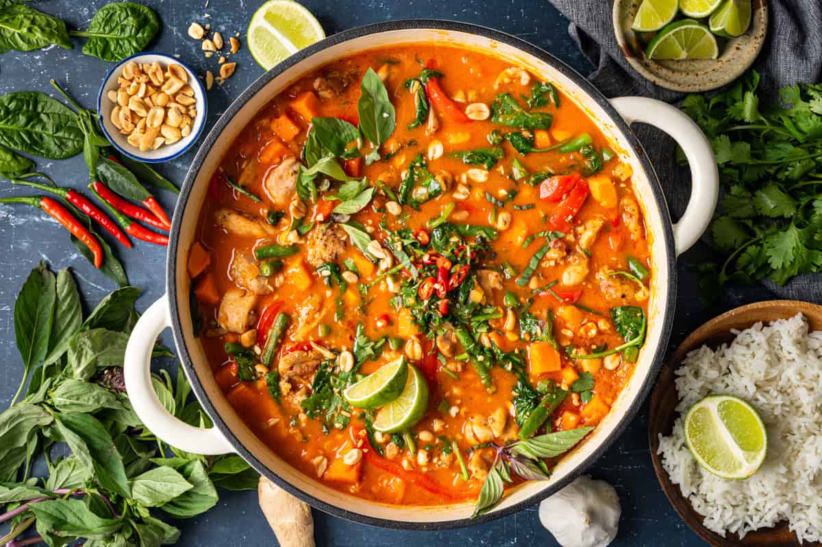 Thai Chicken Curry A pot of Thai red curry with chicken, vegetables, and fresh herbs, garnished with lime wedges, peanuts, and chilies. Surrounding the pot are rice, lime, fresh herbs, and various ingredients on a dark surface.