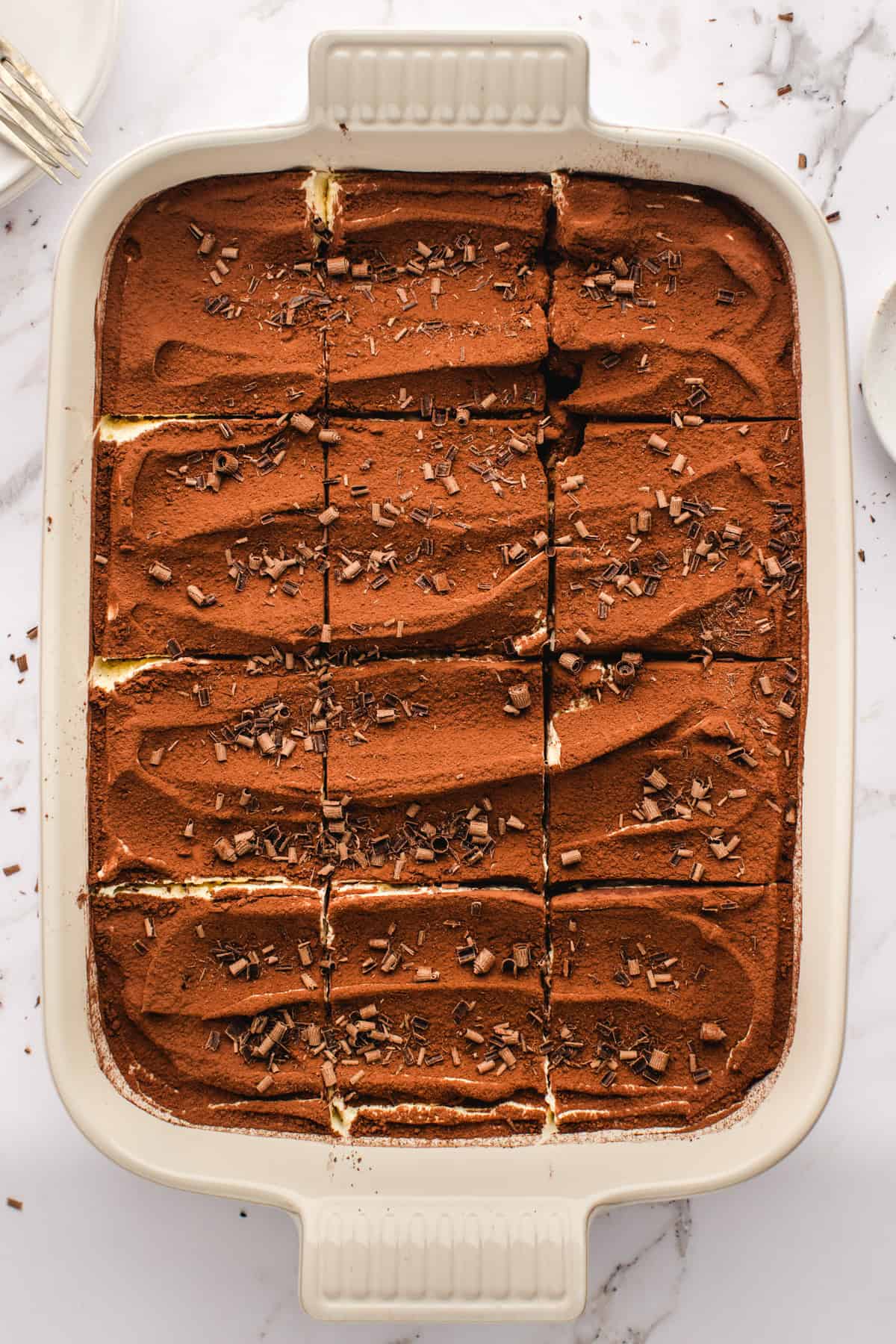 Classic Italian Tiramisu A rectangular baking dish featuring classic tiramisu, cut into neat squares and topped with a layer of cocoa powder and chocolate shavings, rests on a white marble surface.
