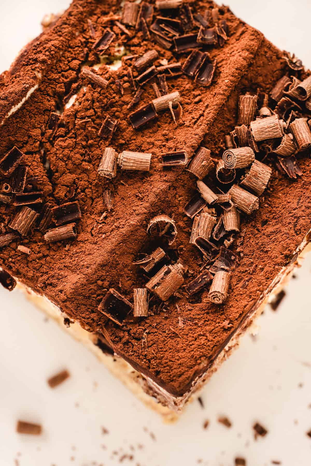 Classic Italian Tiramisu A close-up of a classic tiramisu dessert topped with a layer of cocoa powder and sprinkled with chocolate curls, set on a light-colored surface.