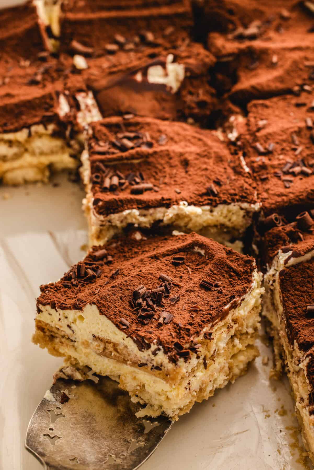 Classic Italian Tiramisu A close-up of classic tiramisu, topped with a dusting of cocoa powder and chocolate shavings, with one piece being lifted on a metal spatula. The creamy, layered dessert sits on a white surface.