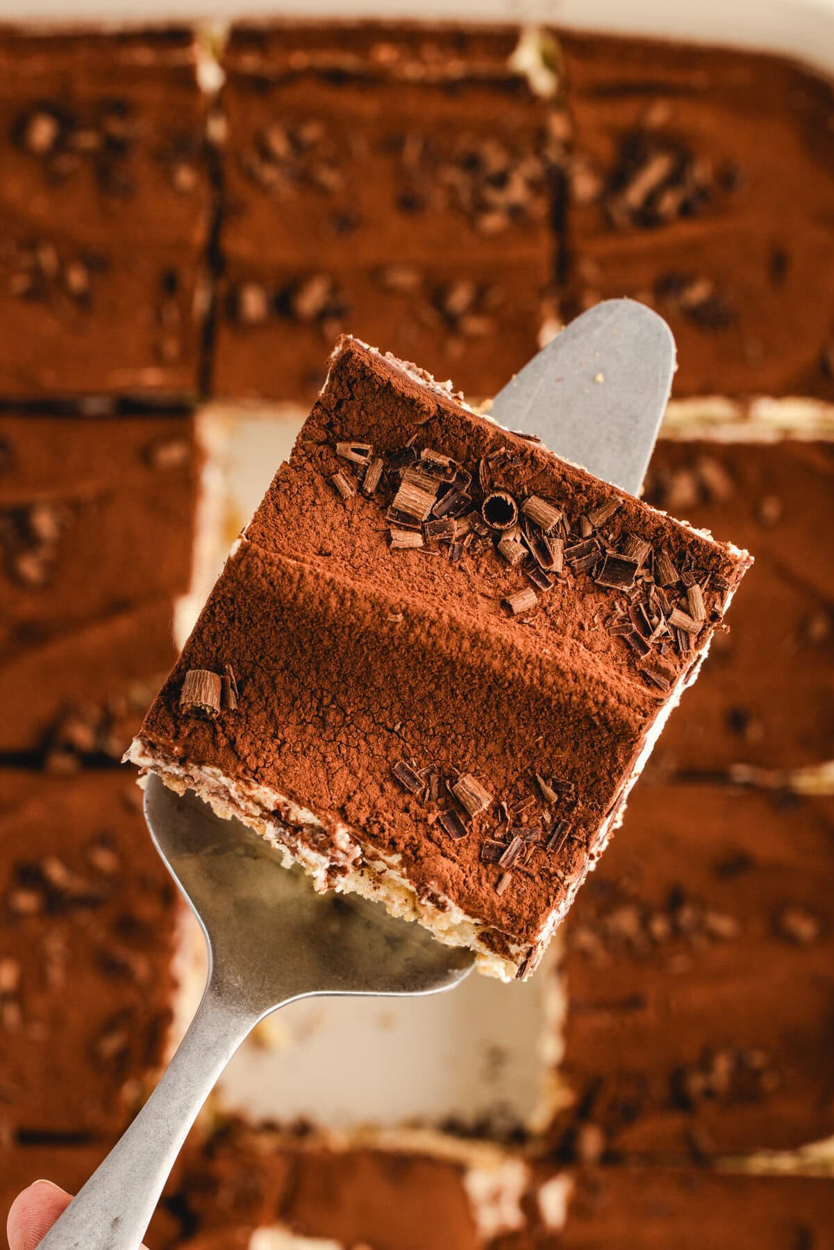 Classic Italian Tiramisu A close-up of a slice of classic tiramisu topped with cocoa powder and chocolate shavings on a serving spatula, with more tiramisu pieces visible in the background.