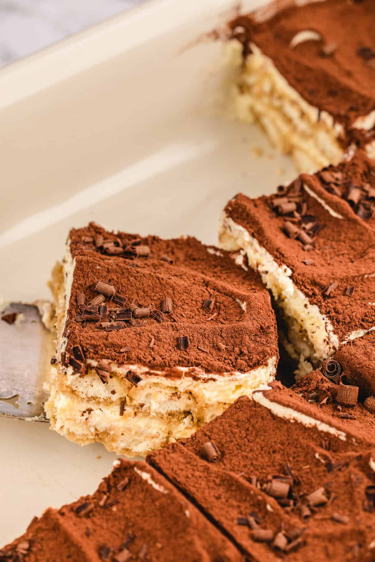 Classic Italian Tiramisu A spatula lifts a square slice of classic tiramisu from a baking dish. The dessert features layers of cream and biscuits, topped with cocoa powder and chocolate shavings, while other neatly cut pieces remain in the dish.
