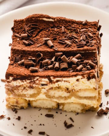 A slice of classic tiramisu on a white plate, topped with cocoa powder and chocolate shavings. The dessert shows creamy layers with ladyfinger biscuits visible at the bottom.