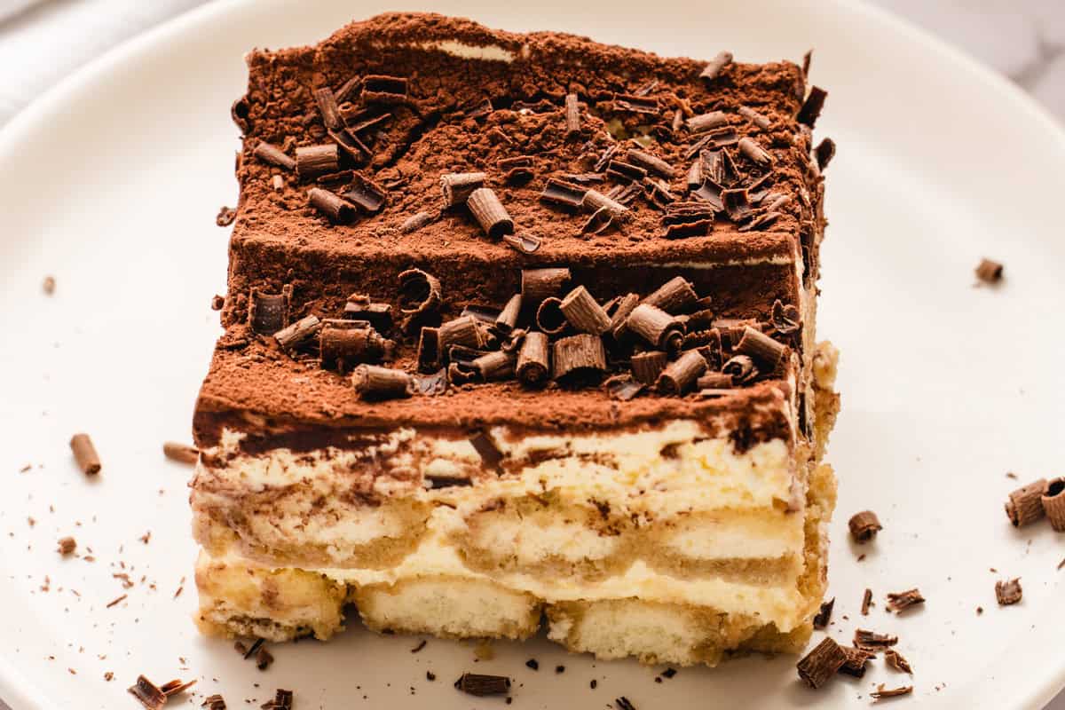 Classic Italian Tiramisu A close-up of a slice of classic tiramisu on a white plate, showing layers of creamy filling and soaked ladyfinger biscuits, topped with cocoa powder and chocolate shavings.