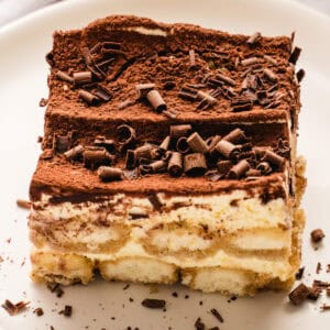 A close-up of a slice of classic tiramisu on a white plate, topped with cocoa powder and chocolate shavings, showing creamy layers and ladyfinger biscuits.