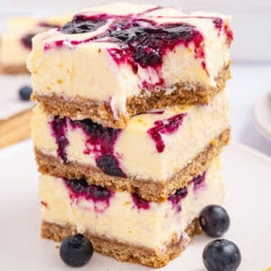 Three blueberry cheesecake bars with a graham cracker crust and a vibrant blueberry swirl are stacked on a plate, with fresh blueberries scattered nearby.