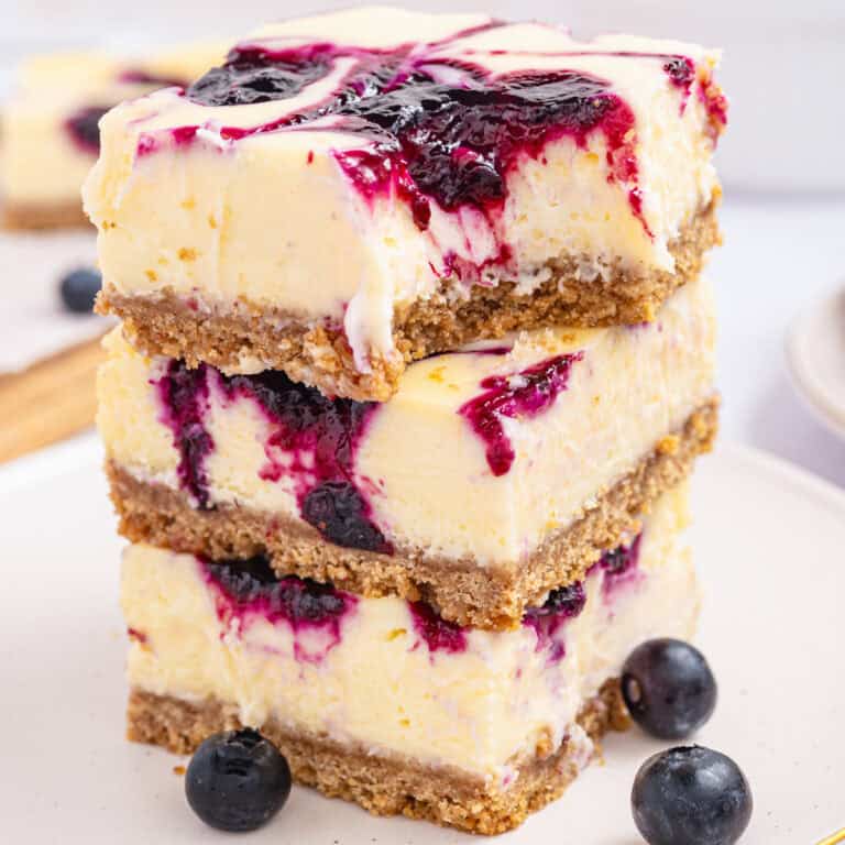 Three blueberry cheesecake bars with a graham cracker crust and a vibrant blueberry swirl are stacked on a plate, with fresh blueberries scattered nearby.
