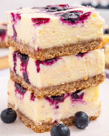 Three blueberry cheesecake bars are stacked on a plate, revealing creamy layers, a crumbly crust, and vibrant blueberry swirls.