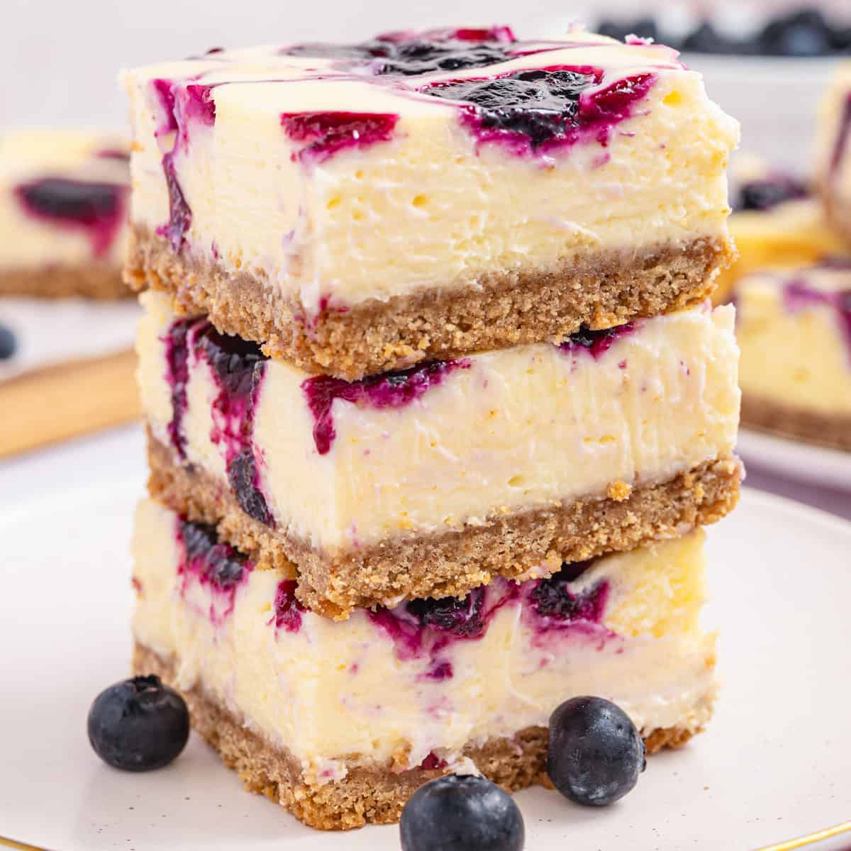 Three blueberry cheesecake bars are stacked on a plate, revealing creamy layers, a crumbly crust, and vibrant blueberry swirls.