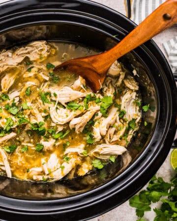 Shredded salsa verde chicken in a slow cooker with green sauce, garnished with fresh cilantro. A wooden spoon stirs the mixture. Fresh cilantro and a cut lime are nearby on the counter.