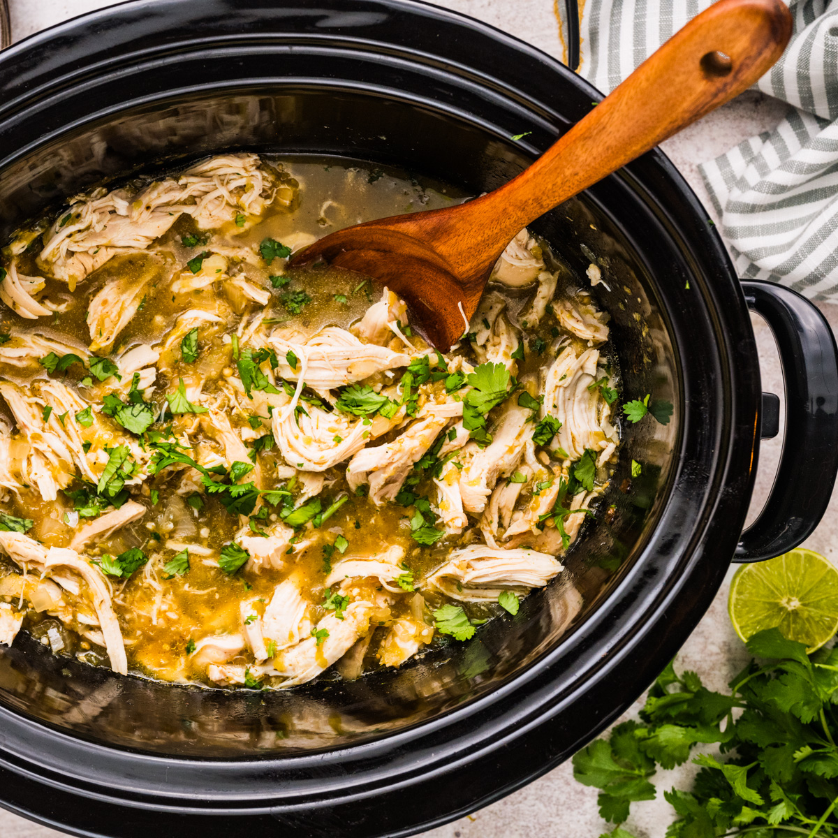 Shredded salsa verde chicken in a slow cooker with green sauce, garnished with fresh cilantro. A wooden spoon stirs the mixture. Fresh cilantro and a cut lime are nearby on the counter.