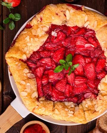 A rustic strawberry galette topped with fresh mint and sliced almonds sits on a white pie dish, surrounded by strawberries, almond slices, and brown sugar on a wooden table.