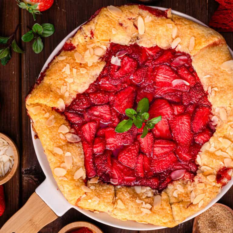 A rustic strawberry galette with a golden, flaky crust, topped with fresh sliced strawberries and almond flakes, garnished with mint leaves-displayed on a white platter on a wooden table.