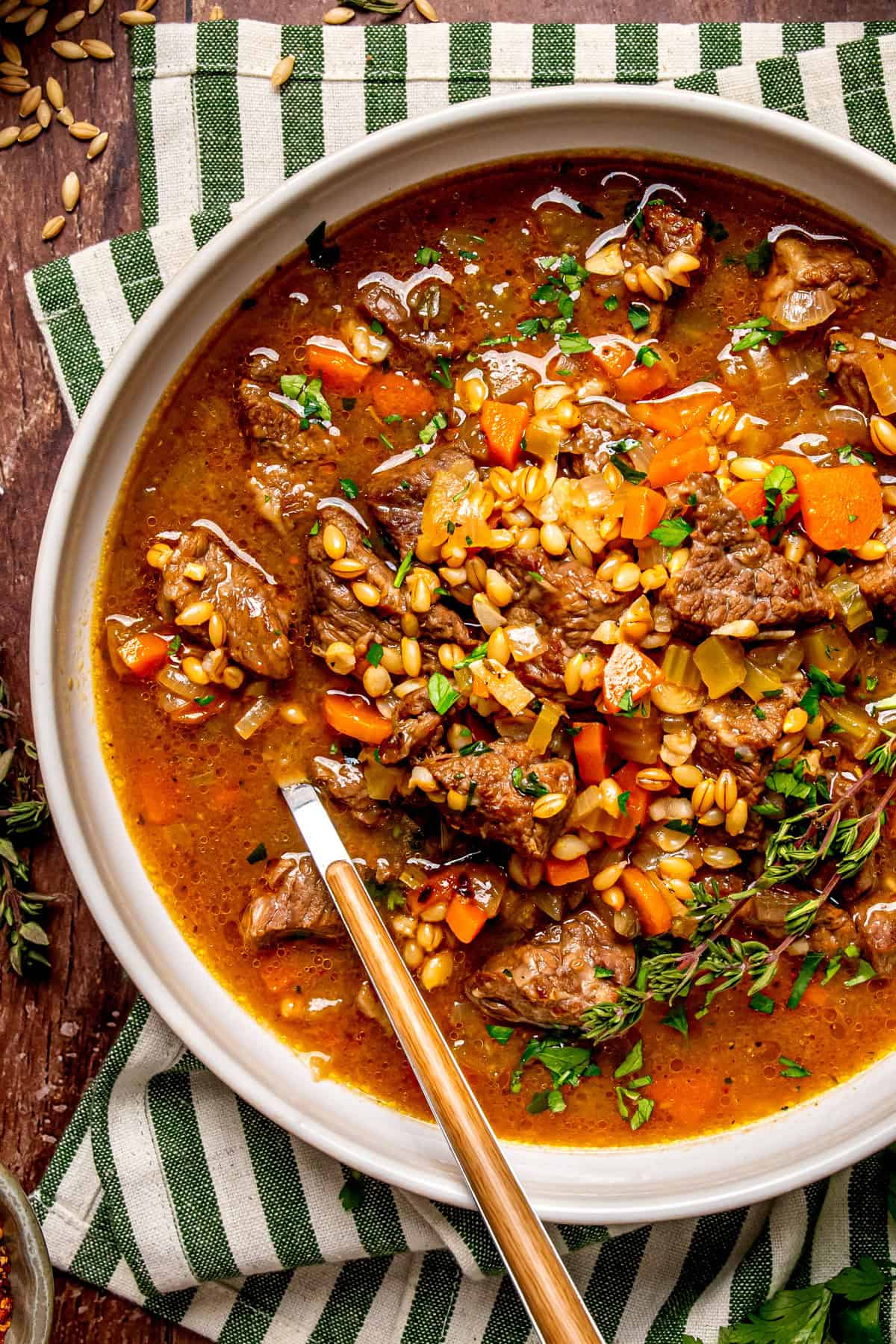 A bowl of hearty beef and vegetable soup with barley, diced carrots, celery, and fresh herbs, served on a striped green and white cloth with a spoon resting inside the bowl.