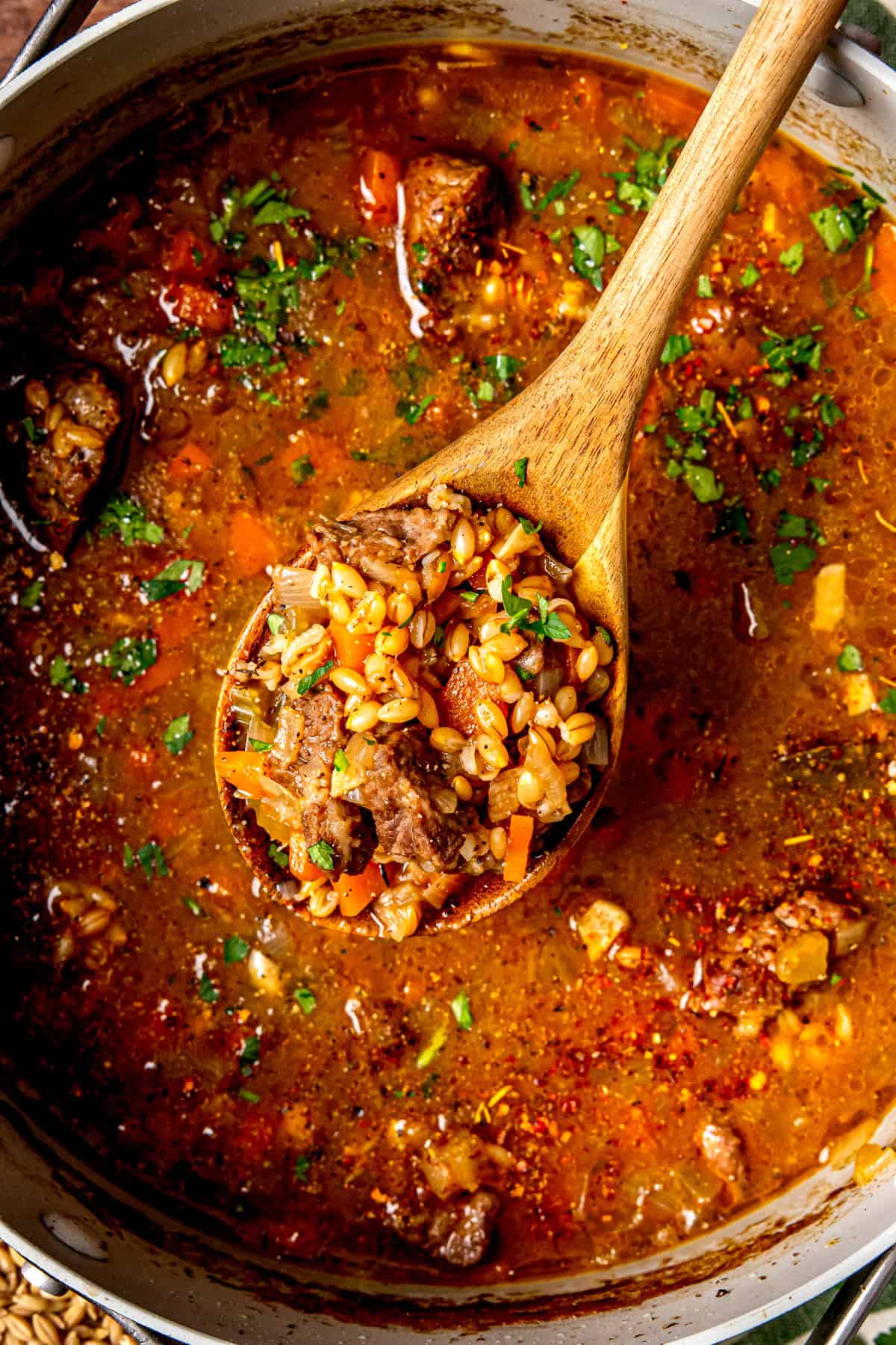 A wooden spoon is lifting a hearty serving of beef and barley soup with visible chunks of beef, barley, carrots, and parsley from a pot filled with rich, reddish broth.