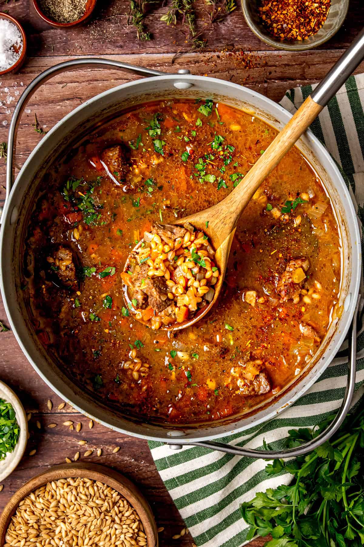 A pot of hearty beef and barley soup sits on a wooden table, with a wooden spoon lifting a serving. The soup is filled with beef chunks, barley, vegetables, and herbs, garnished with chopped parsley.