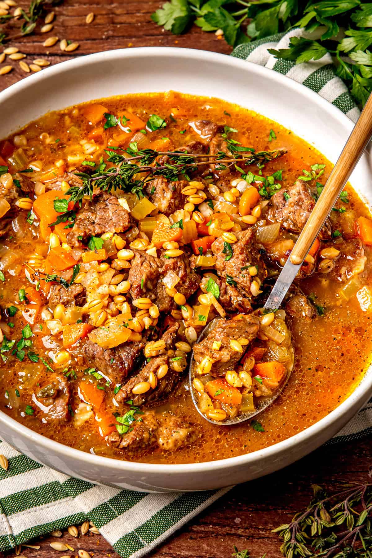 A bowl of hearty beef and vegetable stew garnished with fresh herbs and grains sits on a striped cloth napkin, with a spoon resting inside. The stew contains chunks of beef, diced vegetables, and a rich, brown broth.